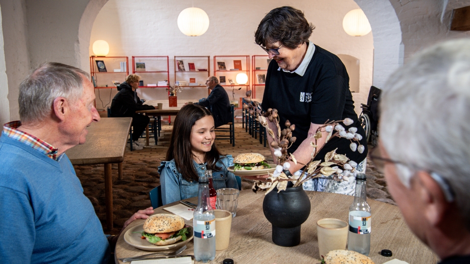 Lille pige får serveret en sandwich af en kvinde imens sidder ældre herre og smiler.