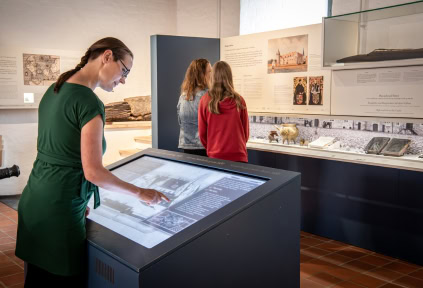 Eine Frau verwendet die digitale Vermittlungsstation. In Hintergrund steht eine Frau mit ihrer Tochter und liest in der Ausstellung von Schloss Sonderburg