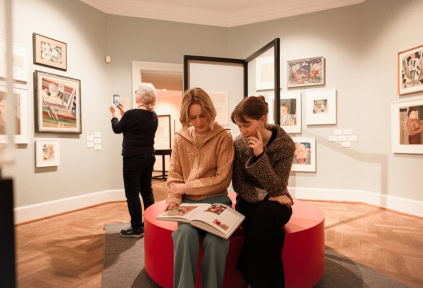 Zwei junge Frauen lesen ein Buch in der Ausstellung, und eine Frau macht ein Foto von einem Kunstwerk.
